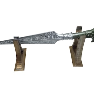 Pode incluir: Uma lança decorativa prateada com detalhes ornamentados, exibida em dois suportes de madeira. A lança apresenta uma lâmina pontiaguda e um cabo detalhado com um detalhe verde. Os suportes são de cor castanho claro.