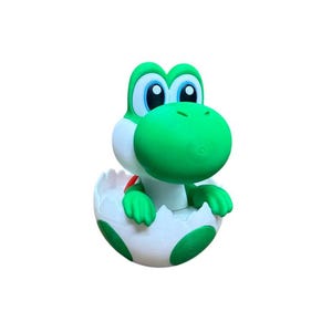 Peut inclure: Figurine verte de Yoshi sortant d'un œuf blanc et vert tacheté. La figurine a de grands yeux bleus, un museau vert et de petits bras. L'œuf est entrouvert, et le fond est blanc uni.