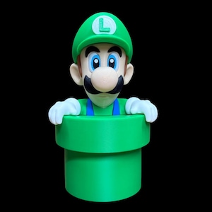 Può includere: Una statuetta di Luigi che emerge da un tubo cilindrico verde. Il personaggio indossa un berretto verde con una lettera L, una salopette blu e guanti bianchi. La figura ha un baffo nero, grandi occhi azzurri e un incarnato chiaro.