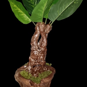 Pode incluir: Uma planta artificial decorativa com grandes folhas verdes e um tronco castanho texturizado. A planta está num vaso castanho de aspeto rústico com musgo verde na base, sobre um fundo preto.