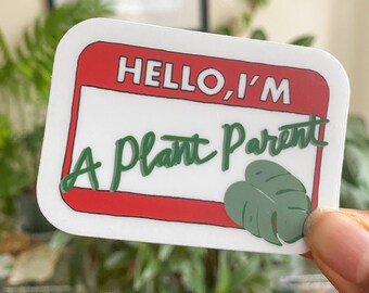 May include: White sticker with a red border and the text "HELLO, I'M A PLANT PARENT" in green cursive writing. A green monstera leaf is in the bottom right corner.