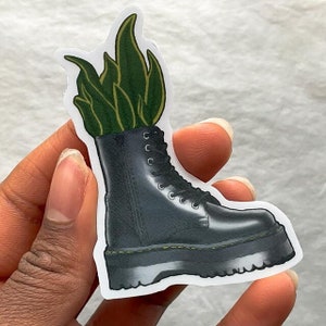 May include: A black leather boot with a green plant growing out of the top. The boot has a thick sole and laces.