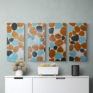 May include: Four rectangular wall art panels with a pebble design in shades of brown, blue, gray, and white. The panels are mounted on a light green wall above a white dresser. A cactus in a white pot and a black speaker are on the dresser.