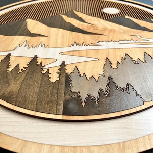 Pode incluir: Uma pe&ccedil;a de arte de parede em madeira com um design de paisagem de montanha. A obra de arte apresenta um efeito em camadas com um rio, &aacute;rvores e montanhas em tons de castanho e cinza. O sol &eacute; vis&iacute;vel no fundo.