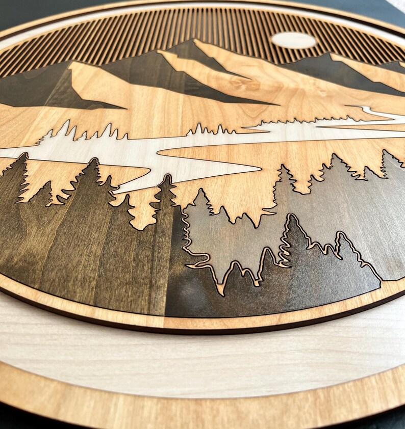 Pode incluir: Uma pe&ccedil;a de arte de parede em madeira com um design de paisagem de montanha. A obra de arte apresenta um efeito em camadas com um rio, &aacute;rvores e montanhas em tons de castanho e cinza. O sol &eacute; vis&iacute;vel no fundo.
