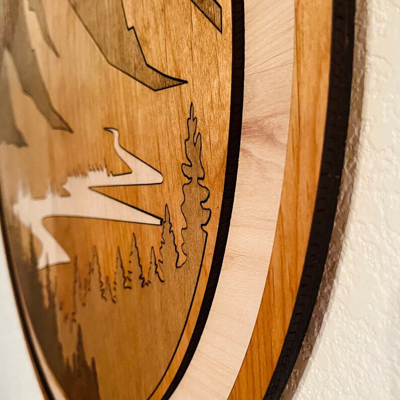 Pode incluir: Arte de parede em madeira com uma cena de montanha e floresta em camadas. A obra de arte utiliza v&aacute;rios tons de madeira castanha e bege, com uma borda preta. O design inclui montanhas, &aacute;rvores e um rio.