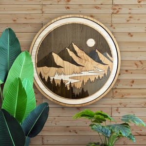 Pode incluir: Uma pe&ccedil;a de arte de parede redonda de madeira com um design de paisagem de montanha. A obra de arte tem uma moldura de madeira clara e um interior detalhado com montanhas, &aacute;rvores, um rio e um sol. A pe&ccedil;a &eacute; exibida contra uma parede de madeira.