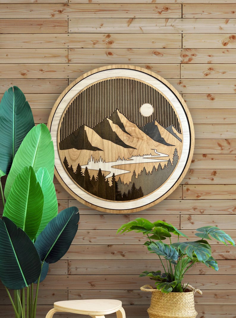 Pode incluir: Uma pe&ccedil;a de arte de parede redonda de madeira com um design de paisagem de montanha. A obra de arte tem uma moldura de madeira clara e um interior detalhado com montanhas, &aacute;rvores, um rio e um sol. A pe&ccedil;a &eacute; exibida contra uma parede de madeira.