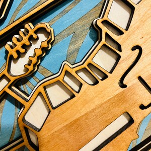 May include: A multi-layered wooden wall art piece featuring a light-colored wood grain finish with dark-edged cutouts. The design includes a stylized guitar headstock and neck silhouette layered over a light blue background with painted wave patterns.