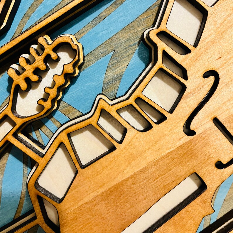 May include: A multi-layered wooden wall art piece featuring a light-colored wood grain finish with dark-edged cutouts. The design includes a stylized guitar headstock and neck silhouette layered over a light blue background with painted wave patterns.