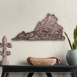May include: A wooden wall art piece in the shape of the state of Virginia. The wood is layered and cut to create a 3D effect. The piece is painted in shades of brown and white.
