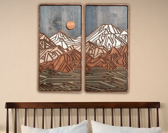 May include: Two-panel wooden wall art depicting a mountain landscape. The artwork features layered wood in shades of brown, white, and green, with a blue-grey sky and a circular sun. The pieces are framed in a dark wood.