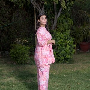 May include: A woman wearing a pink and white floral print two-piece outfit with a long-sleeved top and pants. She is standing in a grassy area with trees in the background.