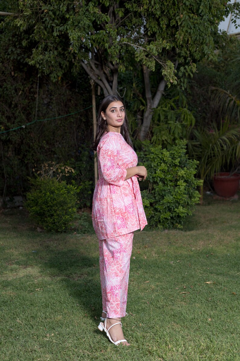 May include: A woman wearing a pink and white floral print two-piece outfit with a long-sleeved top and pants. She is standing in a grassy area with trees in the background.