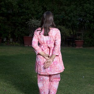 May include: A woman wearing a pink floral print pantsuit with a white belt. She is walking on a grassy lawn.