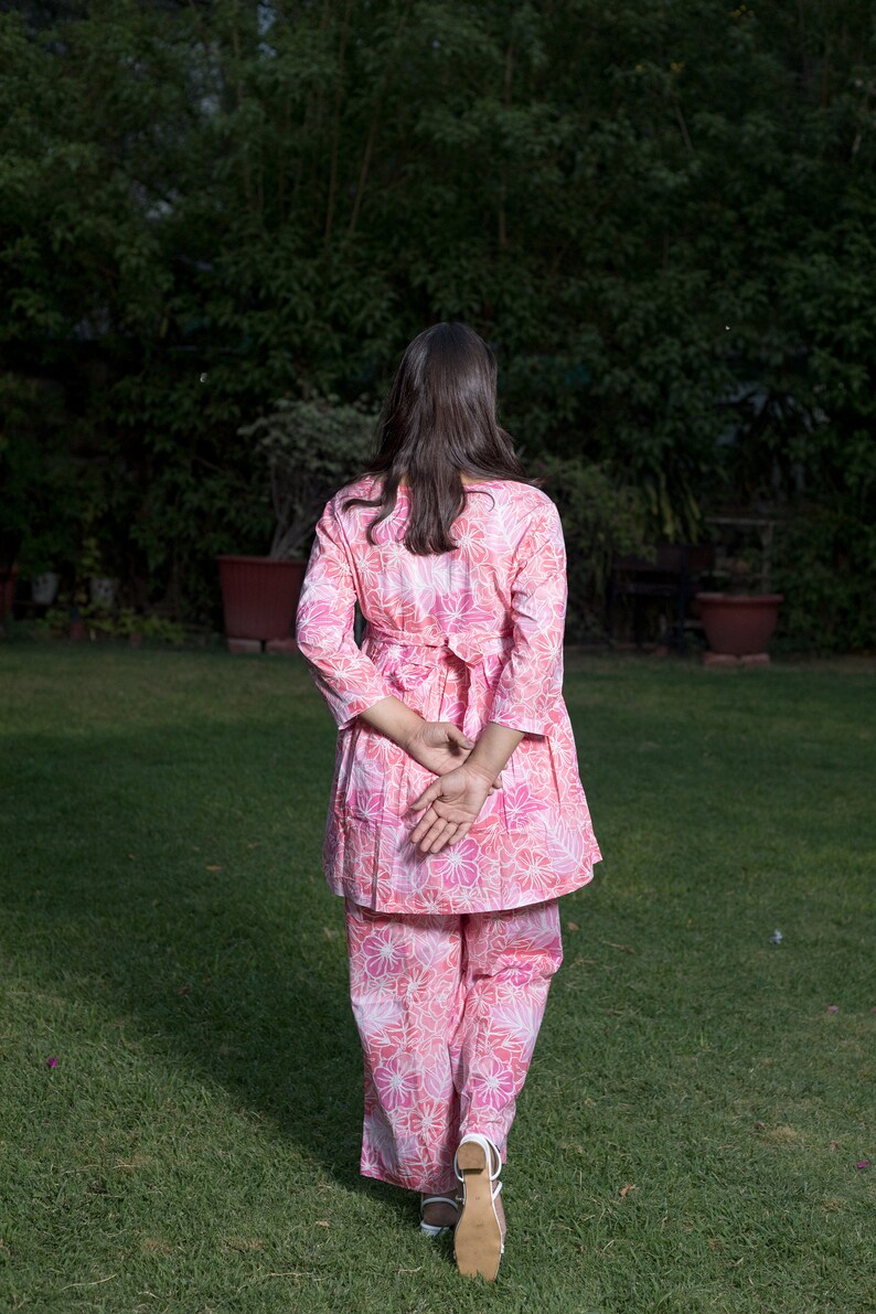 May include: A woman wearing a pink floral print pantsuit with a white belt. She is walking on a grassy lawn.
