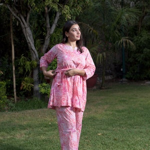 May include: A pink and white floral print two-piece outfit with a long-sleeved top and pants. The top has a tie at the waist.