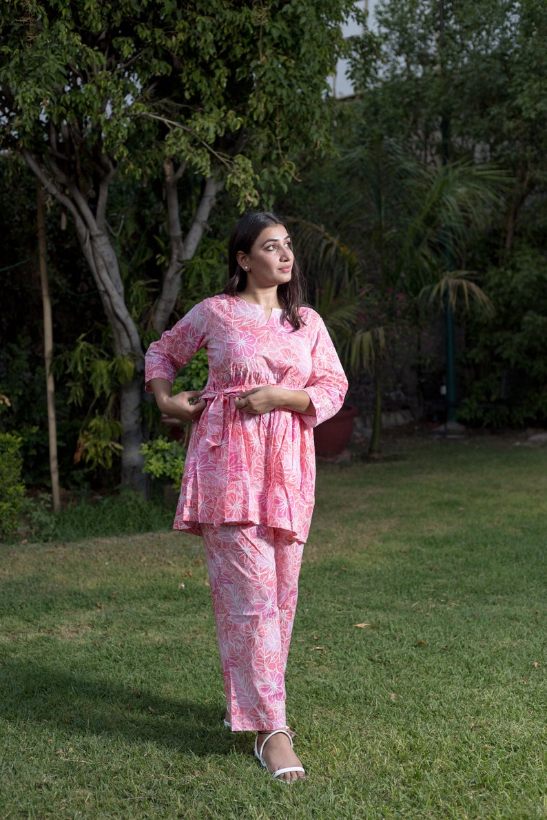 May include: A pink and white floral print two-piece outfit with a long-sleeved top and pants. The top has a tie at the waist.