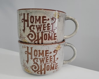 May include: Two stacked ceramic mugs with a speckled brown and white glaze. The mugs feature the text "Home Sweet Home" in brown lettering with a decorative floral design.
