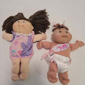 May include: Two Cabbage Patch Kids dolls. One doll has brown yarn hair, blue eyes, and wears a pink and purple romper. The other doll has brown hair, a pink sash that says "Happy 2000", and a white diaper.
