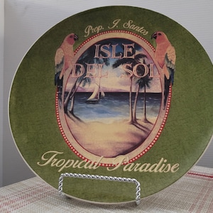 May include: Green ceramic plate with a tropical scene featuring palm trees, a sailboat, and two parrots. The plate has a red and white border with the text "Isle Del Sol" and "Tropical Paradise".