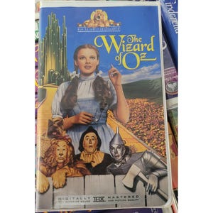 May include: A VHS tape of "The Wizard of Oz" movie. The cover features Judy Garland as Dorothy, along with the Tin Man, Scarecrow, Lion, and Toto. The title is in yellow, with the MGM logo at the top. The tape is labeled "Digitally Mastered" and "THX".