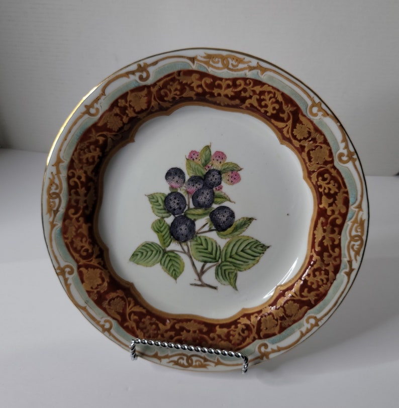 Puede incluir: Un plato de porcelana blanca con un borde dorado y un dise&ntilde;o floral marr&oacute;n y dorado. El centro del plato presenta una imagen pintada a mano de una rama de moras con hojas verdes y moras negras.