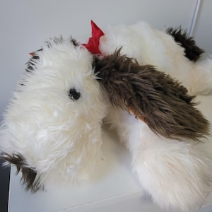 May include: A plush dog toy with white fur, brown ears, and a red bow. The toy has a black eye and is lying down. The dog toy is soft and fluffy, and is a great gift for children.