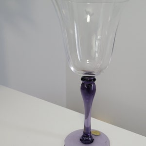 May include: A clear glass wine glass with a purple stem and base. The glass has a wide bowl and a delicate, fluted design.