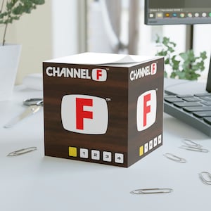 May include: A brown wooden desk organizer with a white top and a red "F" on the front. The organizer has the text "CHANNEL F" on the front and the numbers "1 2 3 4" on the bottom.