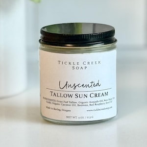 Peut inclure: Un pot en verre de crème solaire Unscented Tallow de Tickle Creek Soap avec un couvercle noir. L'étiquette indique les ingrédients et le poids net de 113 g. Le pot est sur une surface blanche.
