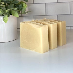 May include: Three bars of natural soap, stacked on top of each other. The soap is a light beige color and has a smooth, matte finish.