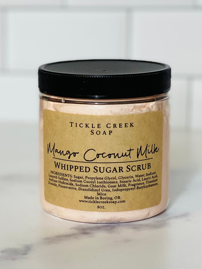 May include: A brown jar with a black lid containing a pink whipped sugar scrub. The label reads "Tickle Creek Soap Mango Coconut Milk Whipped Sugar Scrub" and lists the ingredients. The jar is 8 oz.