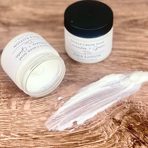 May include: Two white jars of face lotion with black lids. The jars are labelled "Tickle Creek Soap Lavender + Green Tea Face Lotion". One jar is open and a dollop of lotion is on a wooden surface.