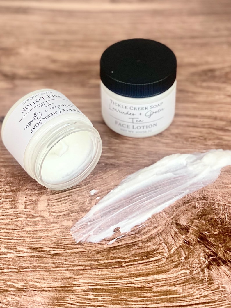May include: Two white jars of face lotion with black lids. The jars are labelled "Tickle Creek Soap Lavender + Green Tea Face Lotion". One jar is open and a dollop of lotion is on a wooden surface.