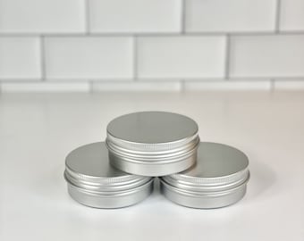 May include: Three small, silver-coloured, round metal tins with ribbed sides and flat lids are arranged in a triangular formation. The tins are empty. The background is a white surface with a white brick wall.