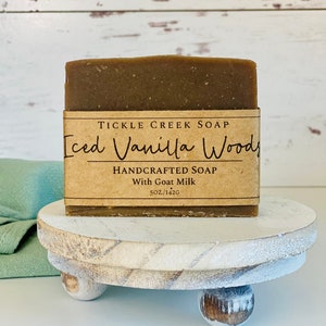 May include: A brown bar of soap with a label that reads "Tickle Creek Soap Iced Vanilla Woods Handcrafted Soap With Goat Milk 5 oz/142g" on a white wooden pedestal.