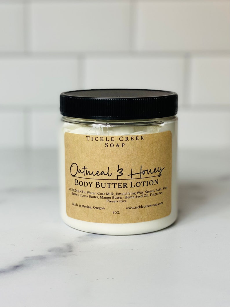May include: A glass jar with a black lid filled with white body butter lotion. The label is brown with black text that reads "Tickle Creek Soap Oatmeal & Honey Body Butter Lotion". The ingredients are listed on the label, along with the website address and the words "Made in Boring, Oregon 8 oz."