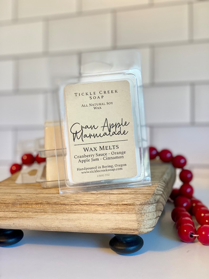 May include: A clear plastic container with a brown label that reads "Tickle Creek Soap, All Natural Soy Wax, Gran Apple Marmalade, Wax Melts, Cranberry Sauce - Orange, Apple Jam - Cinnamon, Handpoured in Boring, Oregon, www.ticklecreeksoap.com, 2.5 oz/72g".