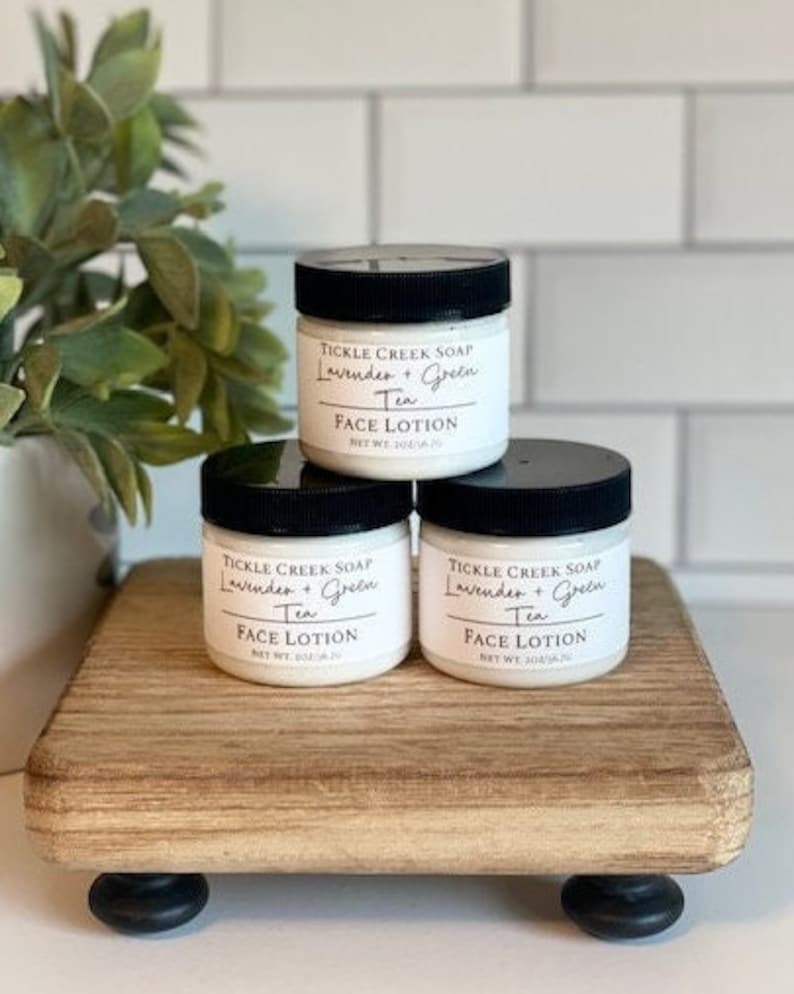 May include: Three white jars of face lotion with black lids and labels that read "Tickle Creek Soap Lavender + Green Tea Face Lotion Net Wt 2 oz/56 g". The jars are sitting on a wooden tray with black feet.