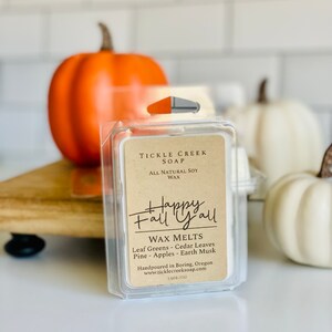 May include: A small, clear plastic container with a brown label that reads "Tickle Creek Soap" and "Happy Fall Y'all". The label also includes the text "Wax Melts" and lists the scents: "Leaf Greens - Cedar Leaves - Pine - Apples - Earth Musk". The container is sitting on a wooden surface with two small pumpkins in the background.