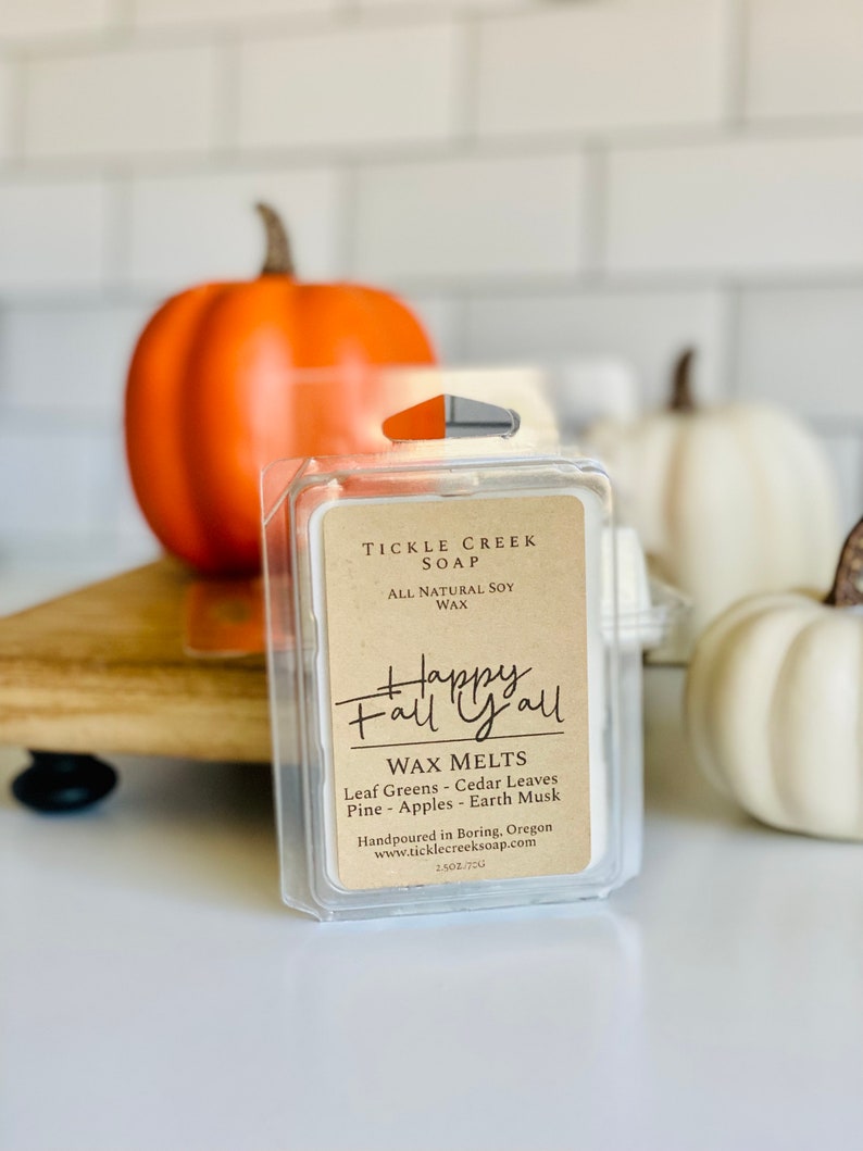 May include: A small, clear plastic container with a brown label that reads "Tickle Creek Soap" and "Happy Fall Y'all". The label also includes the text "Wax Melts" and lists the scents: "Leaf Greens - Cedar Leaves - Pine - Apples - Earth Musk". The container is sitting on a wooden surface with two small pumpkins in the background.