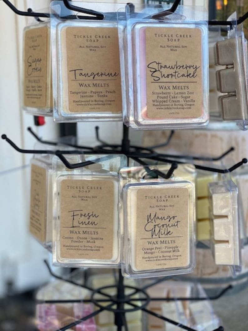 May include: A display of soy wax melts in clear plastic packaging. The wax melts are labeled with the names "Tangerine", "Strawberry Shortcake", "Fresh Linen", and "Mango Coconut Milk". Each label includes the brand name "Tickle Creek Soap" and the text "All Natural Soy Wax".