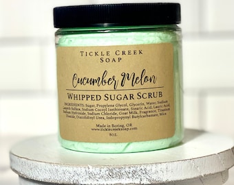 May include: A glass jar with a black lid filled with a green whipped sugar scrub. The label reads "Tickle Creek Soap Cucumber Melon Whipped Sugar Scrub" and lists the ingredients. The jar is sitting on a white wooden pedestal.