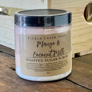 May include: A white jar with a brown label that reads "Tickle Creek Soap Mango & Coconut Milk Whipped Sugar Scrub". The label lists the ingredients and states the size is 8 oz.