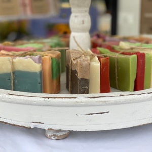 May include: A white wooden tray with a variety of handmade soap bars. The soap bars are layered and tied with twine. The soap bars are in various colors, including green, red, brown, and white.