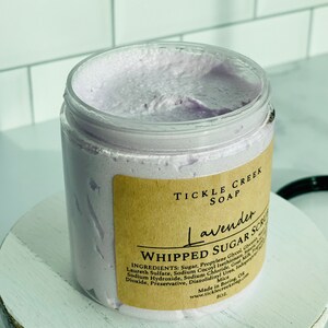 May include: A lavender-coloured whipped sugar scrub in a clear jar with a brown label that reads "Tickle Creek Soap Lavender Whipped Sugar Scrub". The label lists the ingredients: Sugar, Propylene Glycol, Glycerin, Laureth Sulfate, Sodium Cocoyl Isethionate, Sodium Hydroxide, Sodium Chloride, Goat Milk, Fragrance, Mica, Diazolidinyl Urea, Iodopropynyl Butylcarbamate, Preservative. Made in Boring, OK. www.ticklecreeksop.com 8 oz.