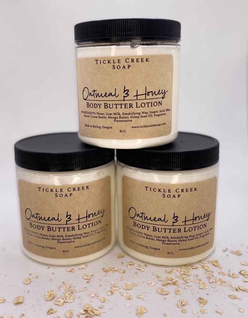 May include: Three white plastic jars with black lids containing Oatmeal & Honey Body Butter Lotion. The jars are labeled with the text "Tickle Creek Soap" and the ingredients list. The jars are sitting on a white surface with scattered oatmeal flakes.