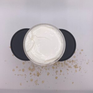May include: A white cream in a clear jar with a black lid. The jar is sitting on a black lid. The cream is surrounded by small, light brown oat flakes.