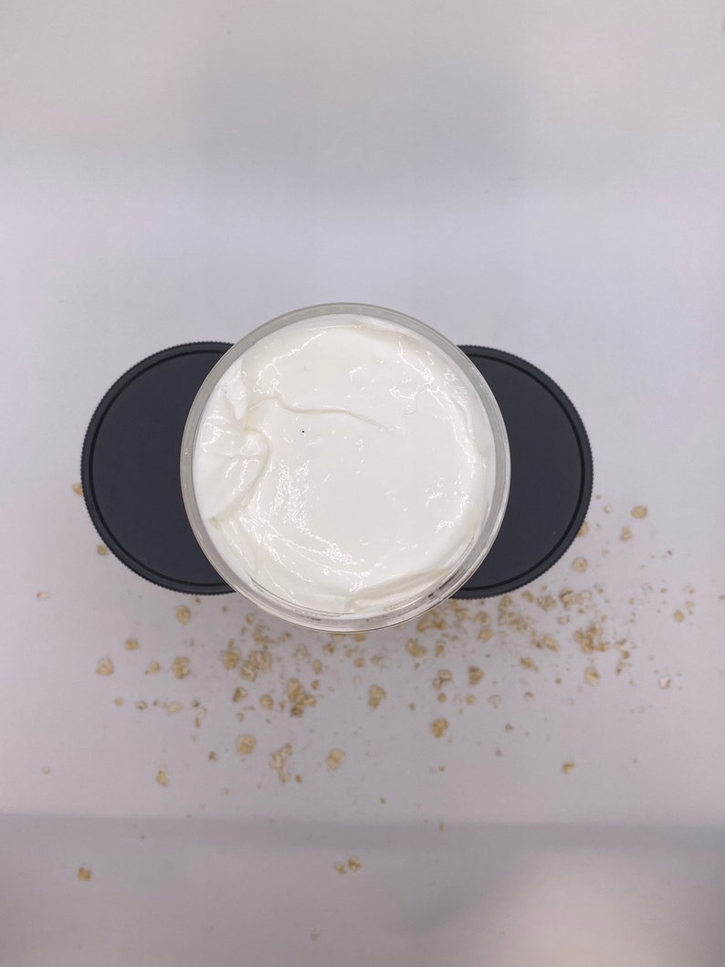 May include: A white cream in a clear jar with a black lid. The jar is sitting on a black lid. The cream is surrounded by small, light brown oat flakes.
